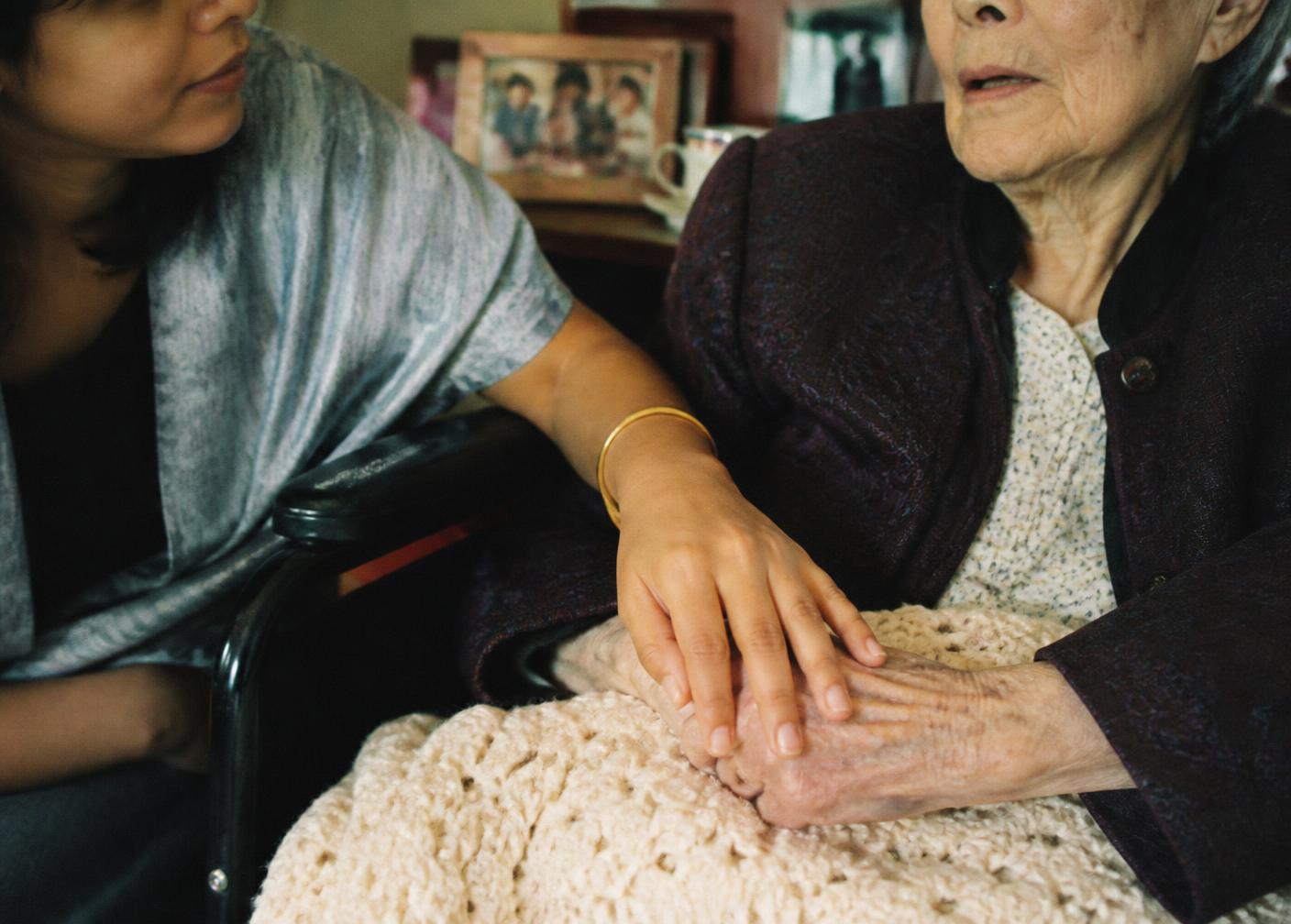 A companion holding the hands of a smiling older woman in a wheelchair